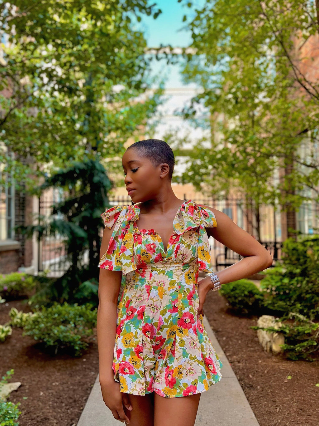 Woman with low hair cut in the middle of a pathway wearing a floral romper with tie straps and a flared pleated bottom