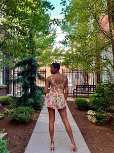 Woman with low hair cut in the middle of a pathway wearing a floral romper with tie straps and a flared pleated bottom. Woman is wearing strappy tie up heels 