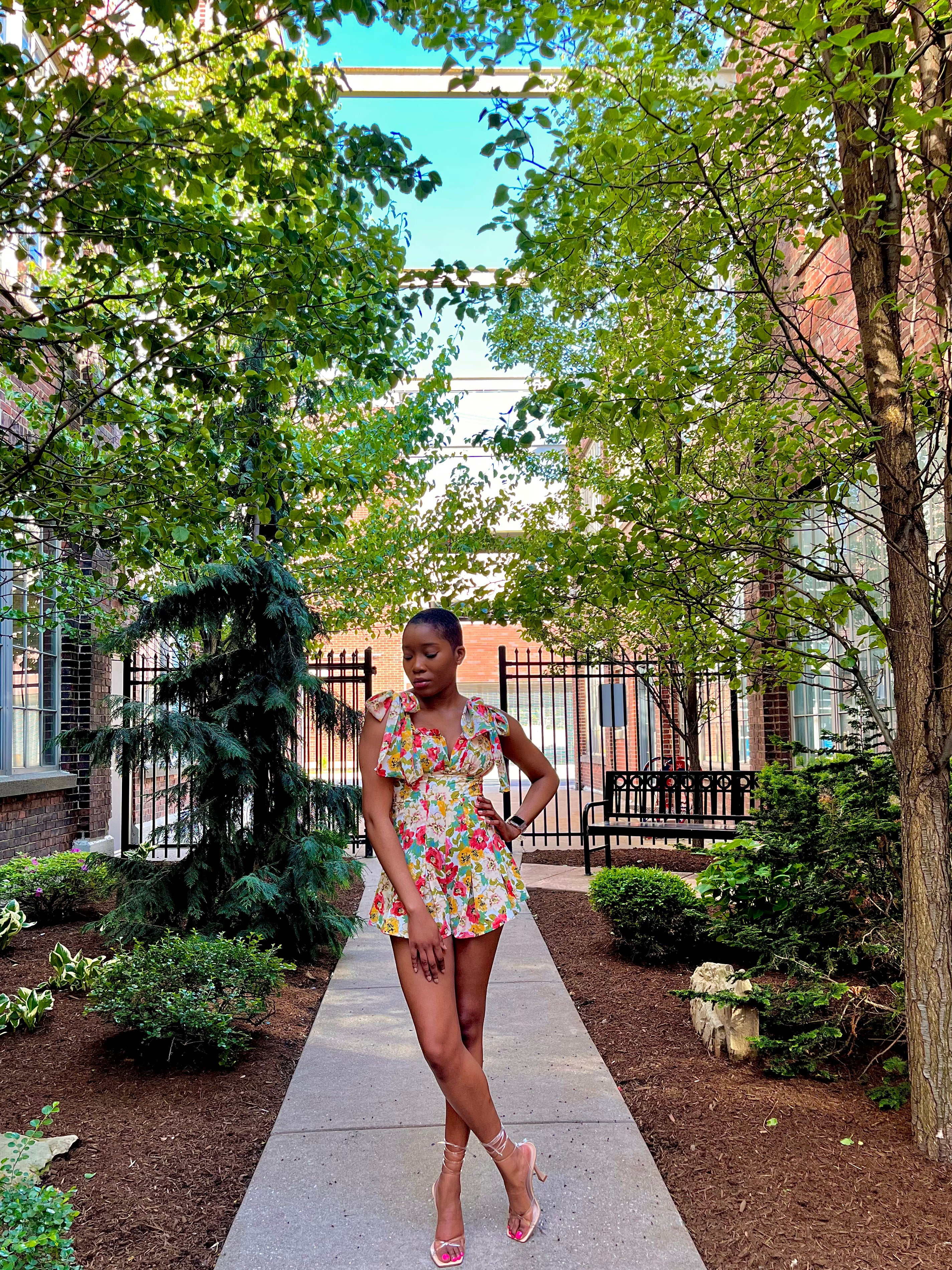 Woman with low hair cut in the middle of a pathway wearing a floral romper with tie straps and a flared pleated bottom. Woman is wearing strappy tie up heels